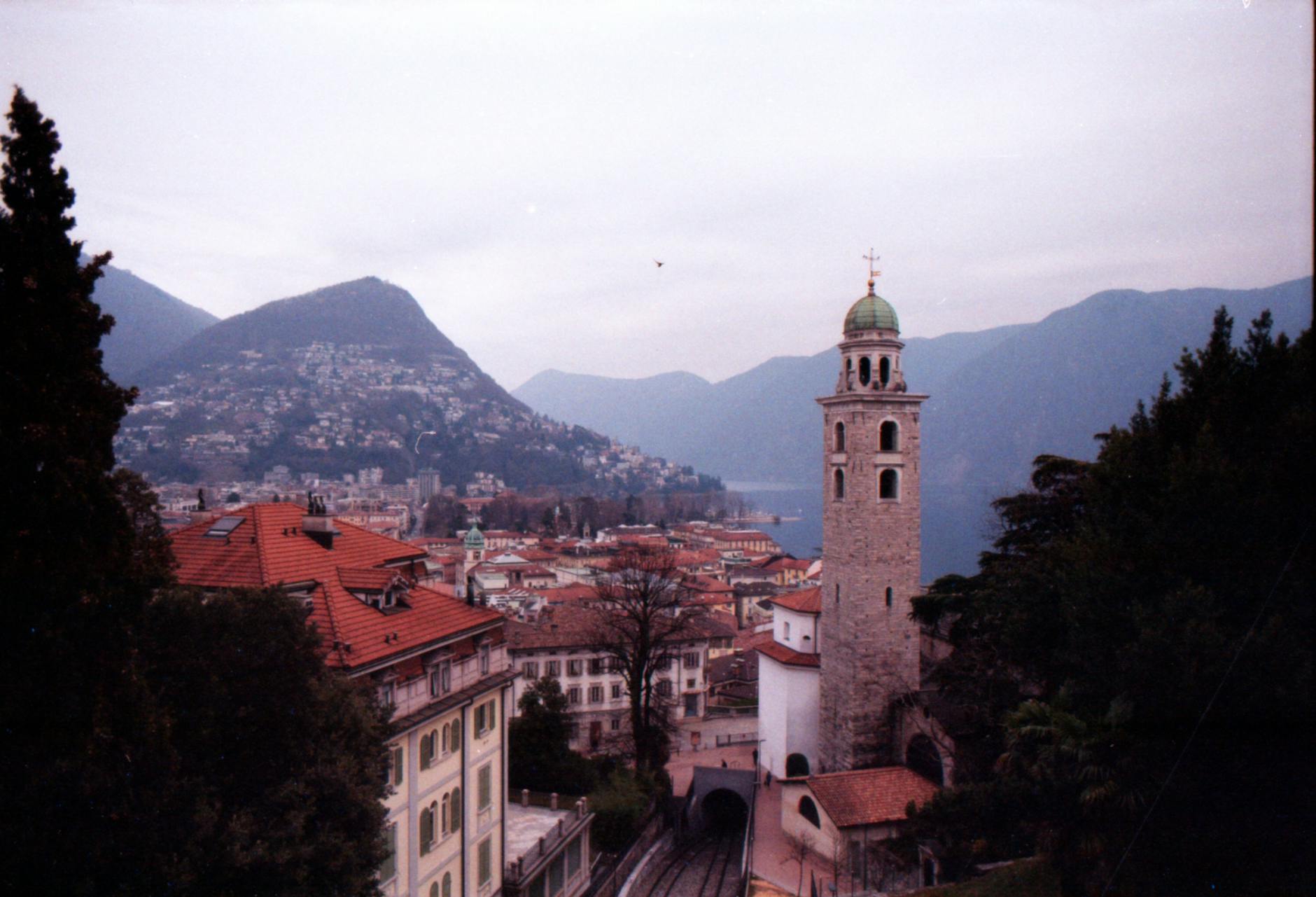 Lugano old town piazza with cafes, palm trees, and lake visible in background, summer afternoon