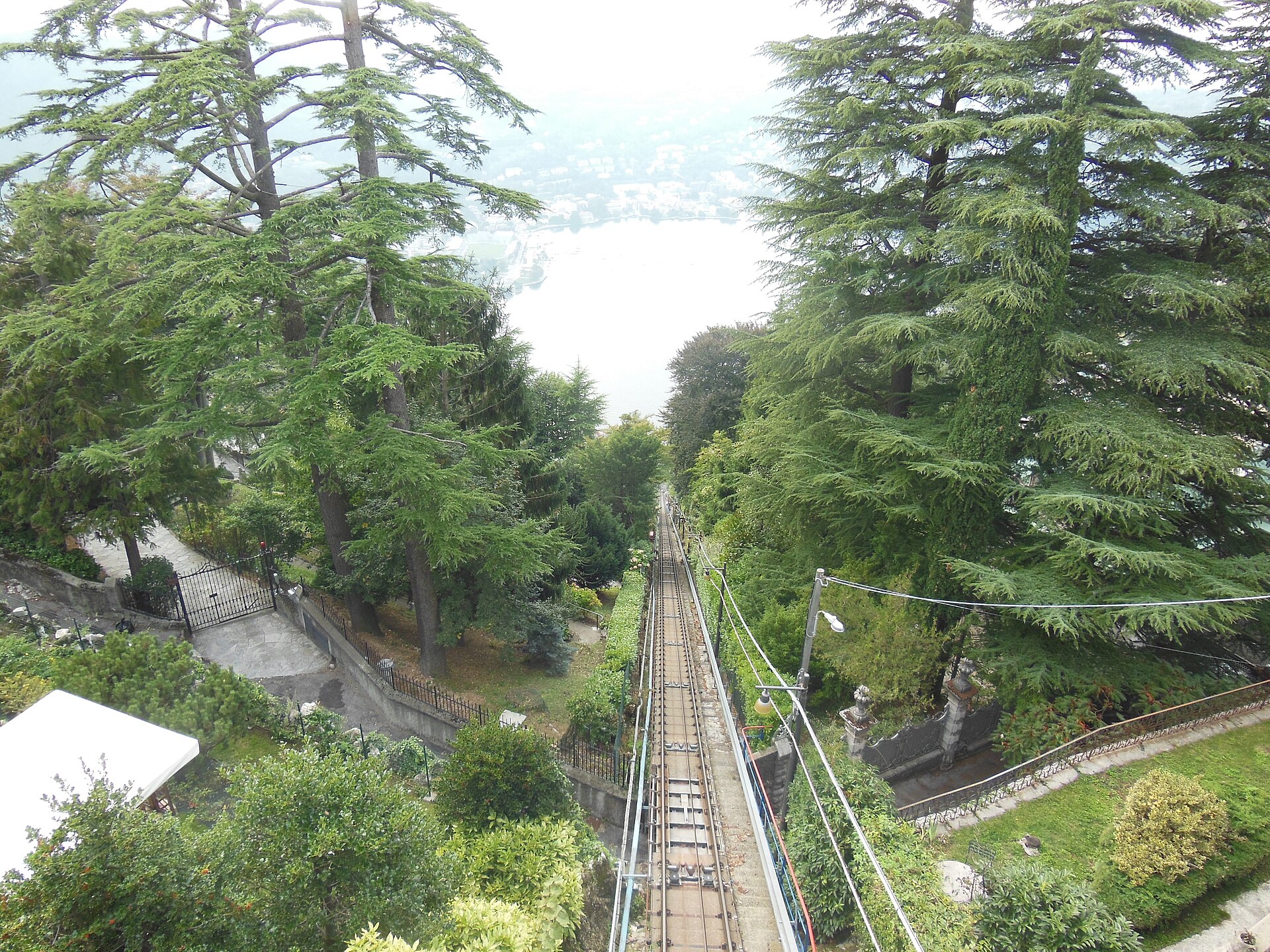 View down the steep single track of the Como to Brunate funicular through pine trees, with Lake Como and the far shore in the haze below