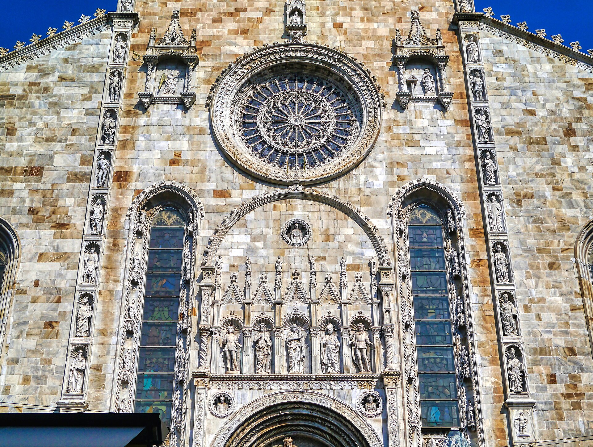 The marble facade of Como Cathedral, showing the rose window, Gothic niches with carved saints and the pointed arch above the main door