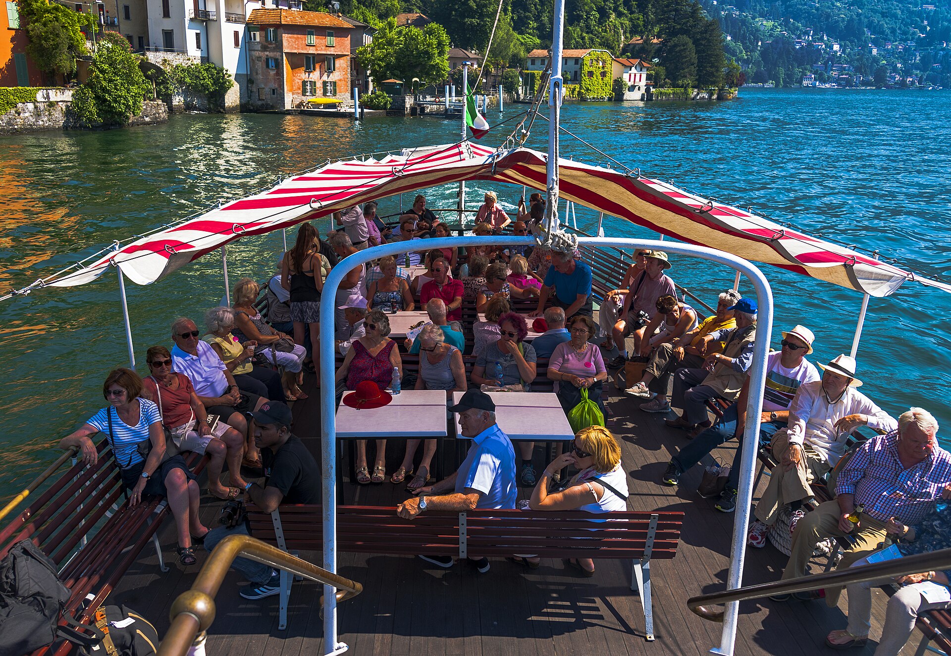 Passengers sitting under a red and white striped awning on the open rear deck of a Lake Como ferry, with a lakeside village and green hillside behind