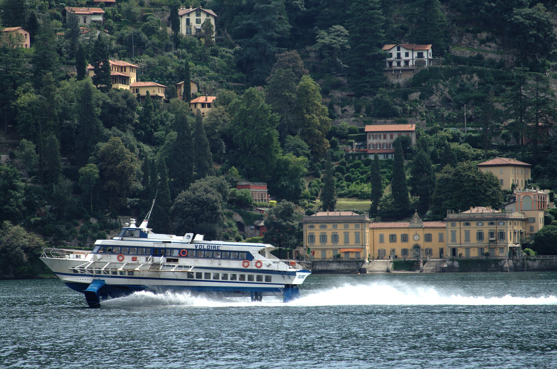 A Navigazione Laghi hydrofoil under way on Lake Como at speed, throwing spray, with villas and a wooded shore behind