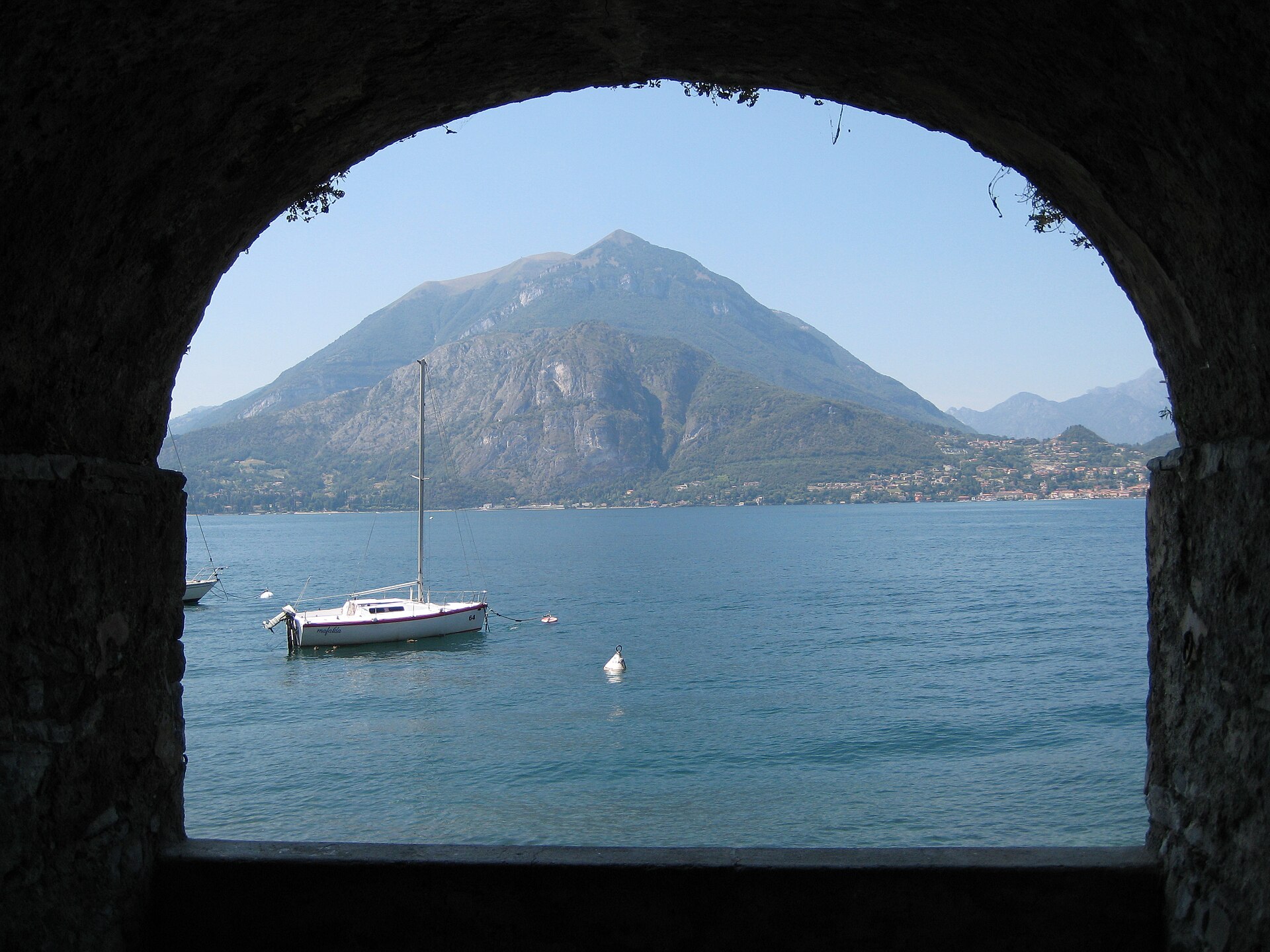 View across Lake Como from a stone archway in Varenna, with a moored sailing boat and a steep mountain rising from the far shore