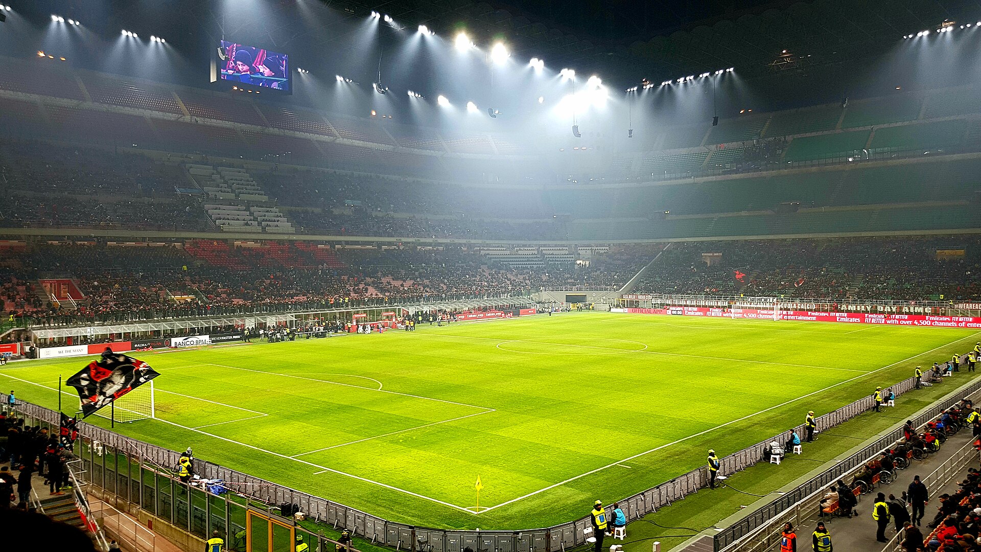 San Siro stadium interior on a match night, floodlit, with the green pitch, three tiers of stands and supporters waving flags behind the goal