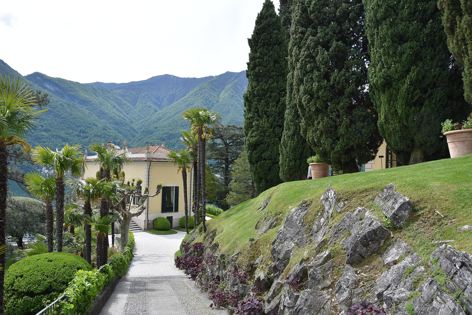 A gravel path curving past palms and clipped box hedges towards the yellow buildings of Villa del Balbianello, with Lake Como and mountains behind